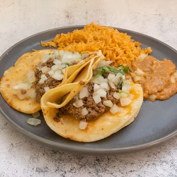 Two El Que Pasa ground beef tacos with onions, rice, and refried beans on a gray plate.